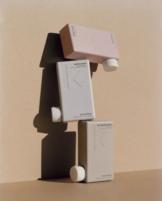 Three Kevin Murphy hair product bottles, 'Angel Wash,' 'Born Again,' and 'Balancing Wash,' are stacked against a light tan background.