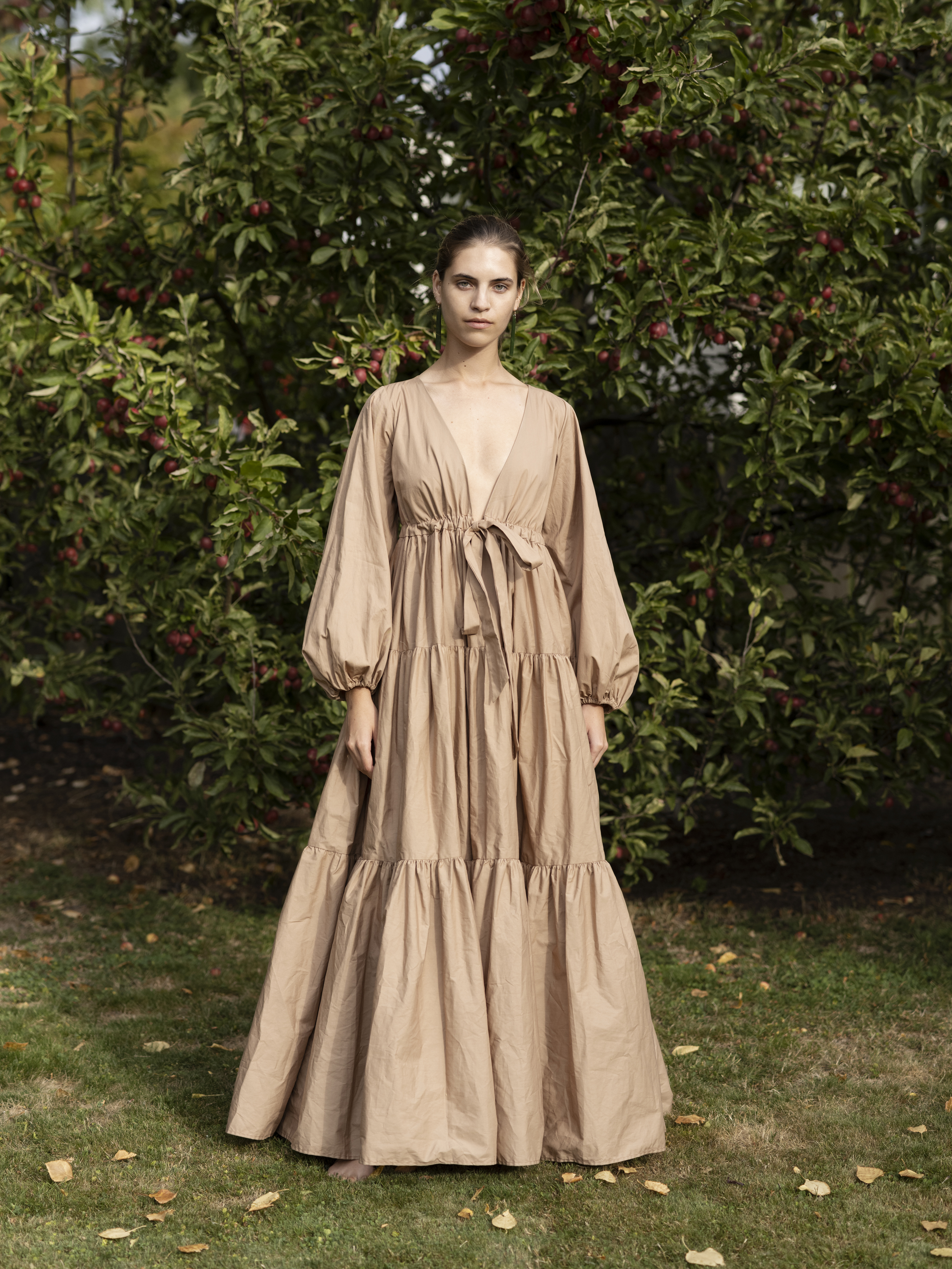 Woman in a long, flowing beige dress stands in a yard with an apple tree; dress features full sleeves and a tie at the waist.