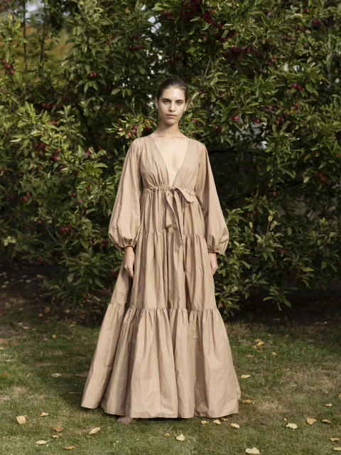 Woman in a long, flowing beige dress stands in a yard with an apple tree; dress features full sleeves and a tie at the waist.