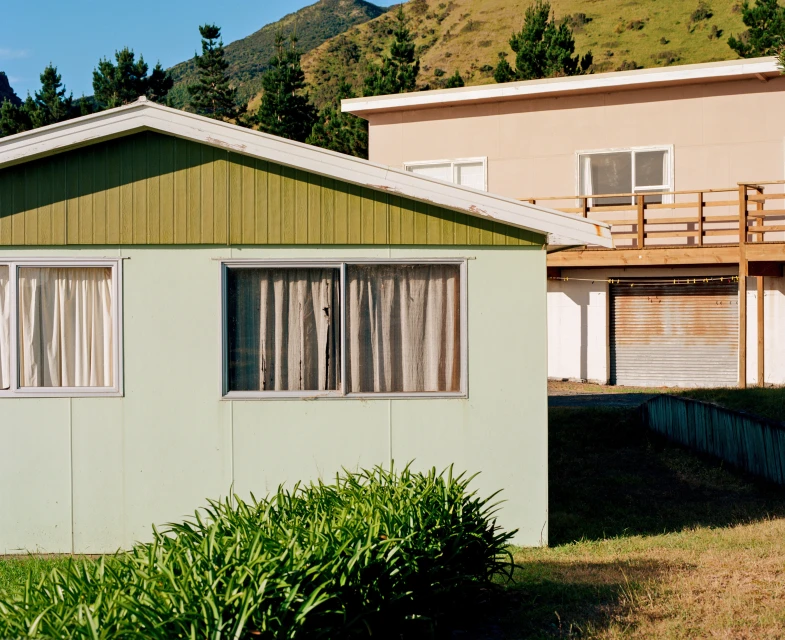 View of a light green house with an olive green roof and trimmed green bushes, with mountains and trees in the background.