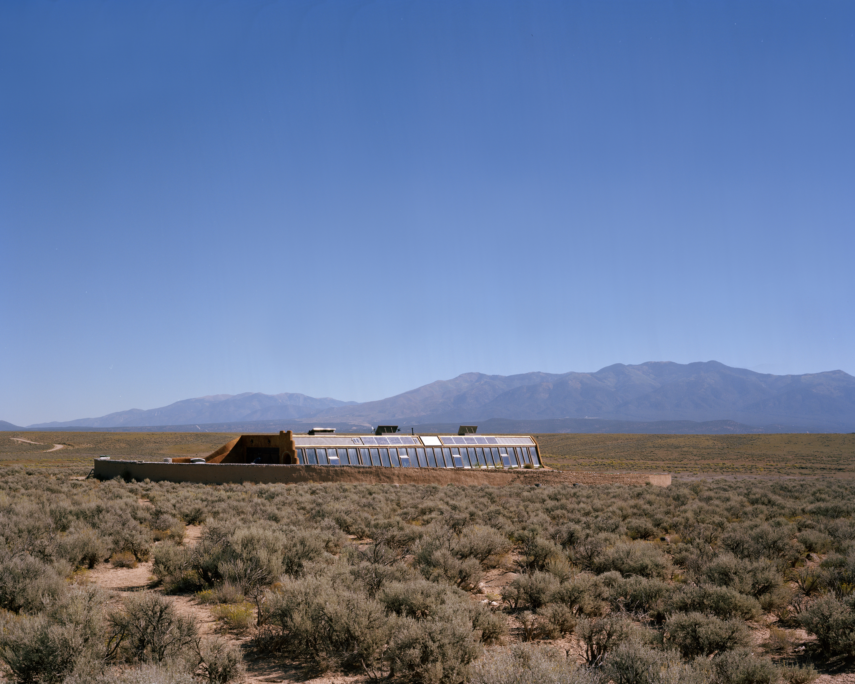 Desert landscape featuring an earthship dwelling with a greenhouse facade, mountains on the horizon, and clear blue sky above.