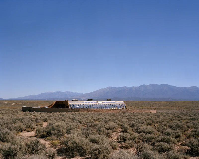 Desert landscape featuring an earthship dwelling with a greenhouse facade, mountains on the horizon, and clear blue sky above.