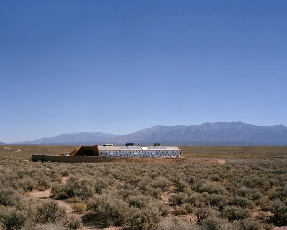 Desert landscape featuring an earthship dwelling with a greenhouse facade, mountains on the horizon, and clear blue sky above.