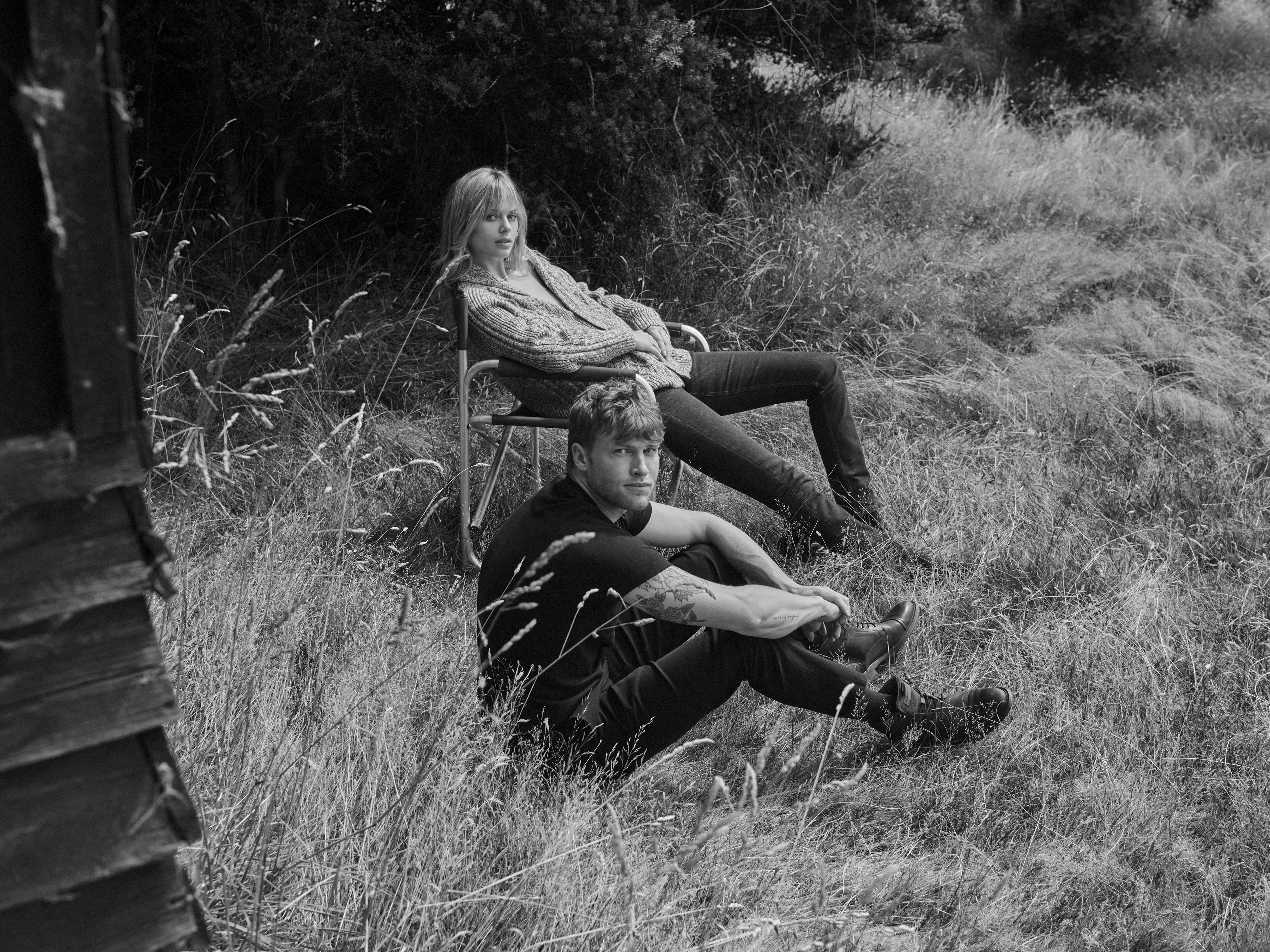 Black and white photo of a young couple outdoors. A woman sits in a chair, while the man sits on the ground in a grassy field.