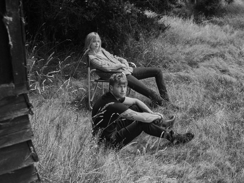 Black and white photo of a young couple outdoors. A woman sits in a chair, while the man sits on the ground in a grassy field.