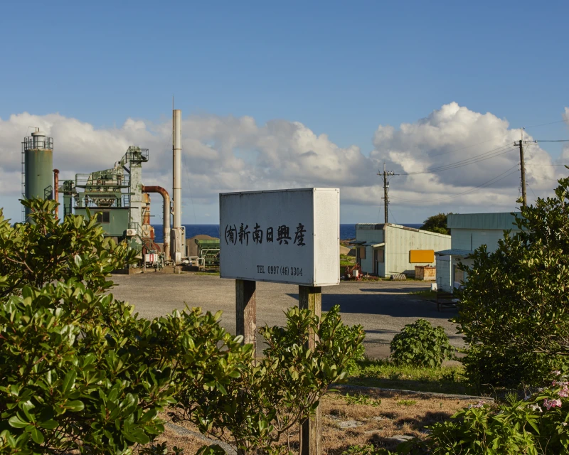 Daytime view of a rustic industrial factory complex with weathered buildings, metal structures, and lush green foliage under a blue sky.