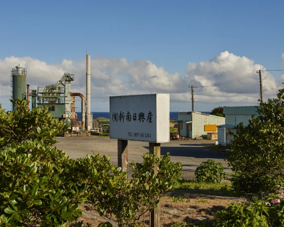 Daytime view of a rustic industrial factory complex with weathered buildings, metal structures, and lush green foliage under a blue sky.