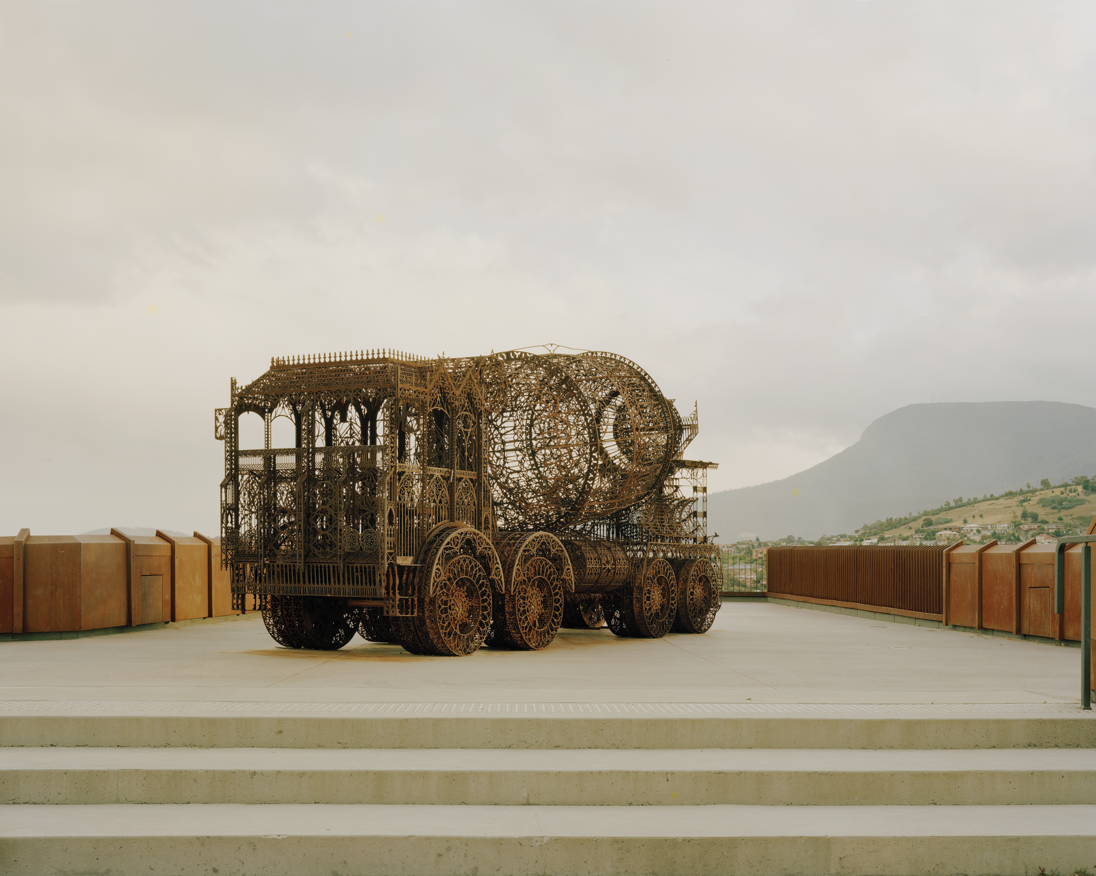 Metal sculpture resembling a semi-trailer truck sits on a concrete platform, set against a backdrop of a cloudy sky and distant mountains.