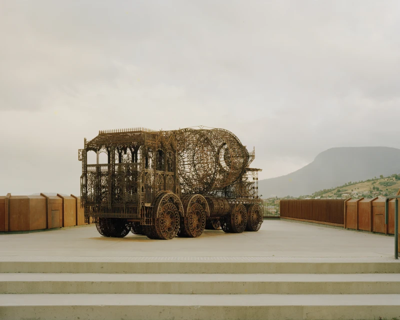 Metal sculpture resembling a semi-trailer truck sits on a concrete platform, set against a backdrop of a cloudy sky and distant mountains.