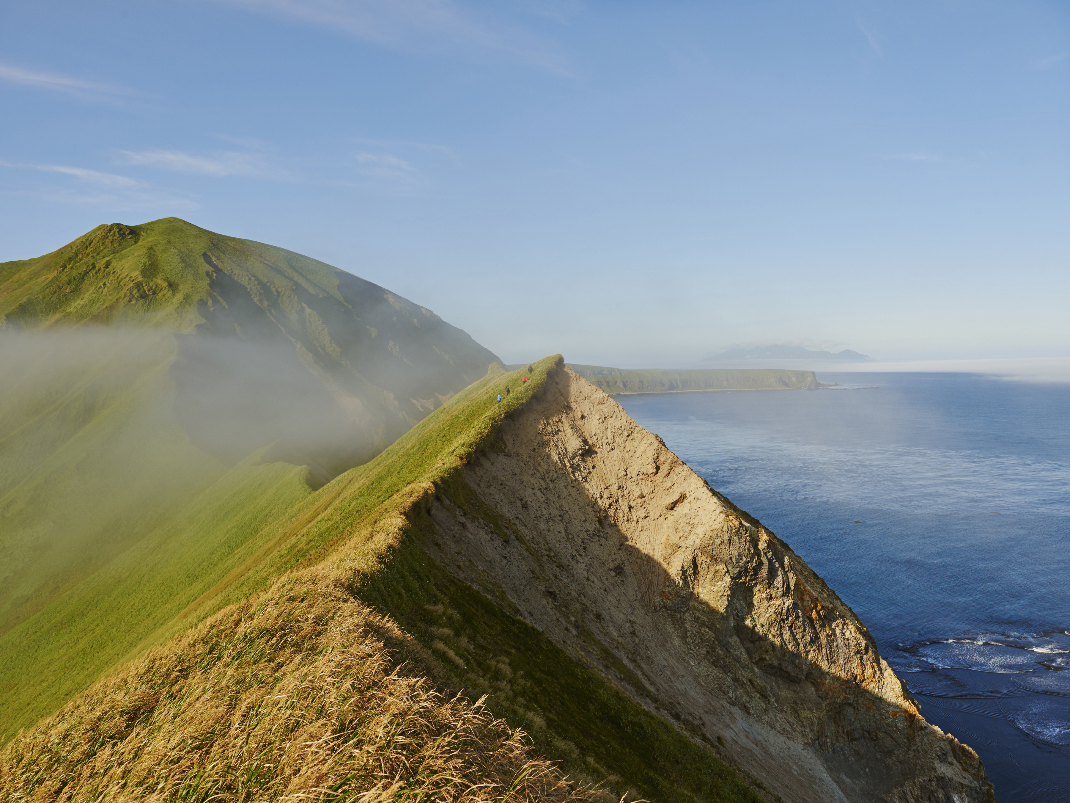 Scenic landscape featuring a grassy mountain ridge meeting the ocean, with a clear blue sky above and mist in the valleys.