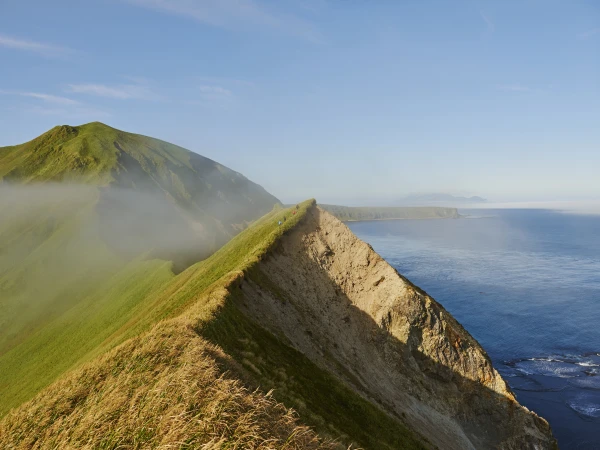 Scenic landscape featuring a grassy mountain ridge meeting the ocean, with a clear blue sky above and mist in the valleys.