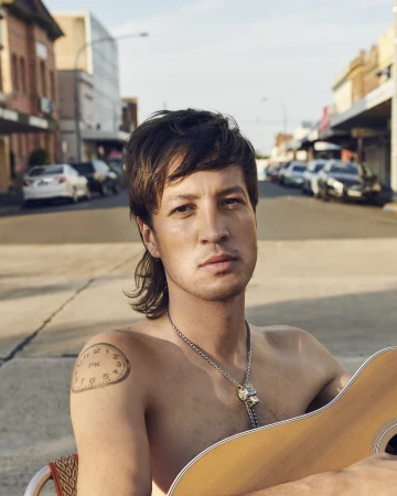 Shirtless man with a mullet and shoulder clock tattoo holding a guitar on a sunny suburban street by Derek Henderson