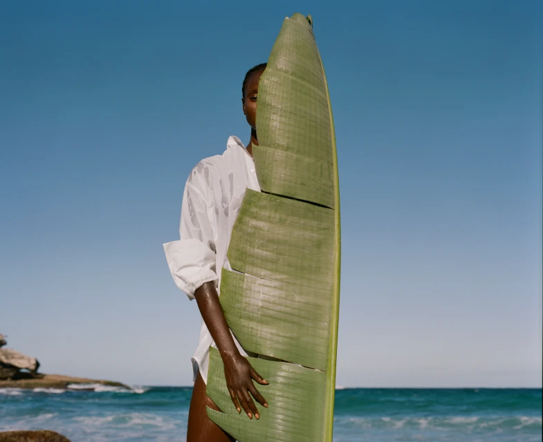 Adult female in a white button-down shirt poses with a large tropical green leaf on a sunny beach with blue skies and ocean.