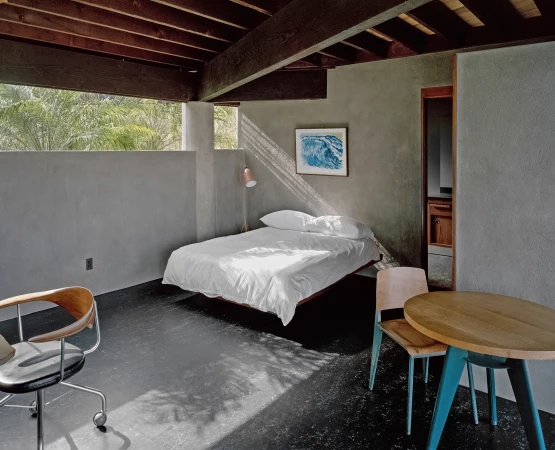 Interior bedroom with a low white bed, exposed wooden ceiling beams, dark floors, and sunlight slanting across the concrete wall.