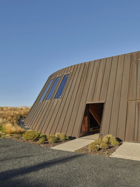 Exterior of a modern building featuring rusty metal facade, slanted windows, and an angular entrance during daytime against a clear blue sky.