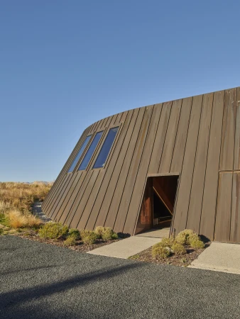 Modern slanted metallic building with angular windows and an open doorway set against sparse landscape and clear blue sky.