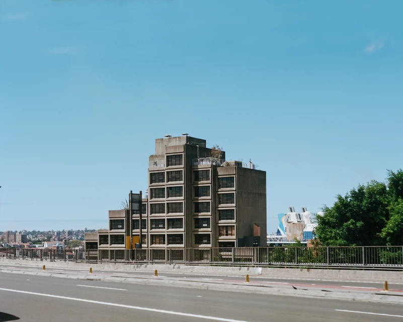 Exterior view of a large concrete building with a flat roof and many windows, situated in an urban environment under a bright blue sky.