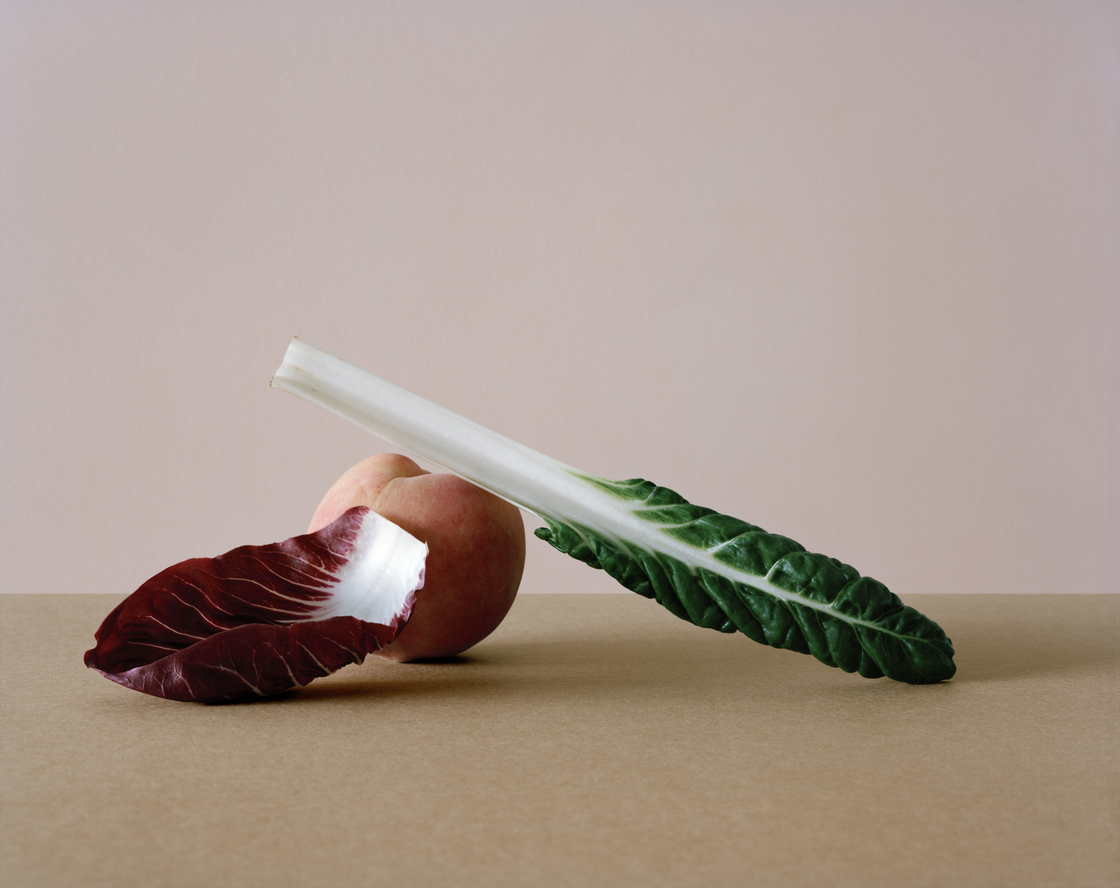Still life photography by Derek Henderson featuring a Swiss chard leaf, a peach, and a radicchio leaf on a tan surface.