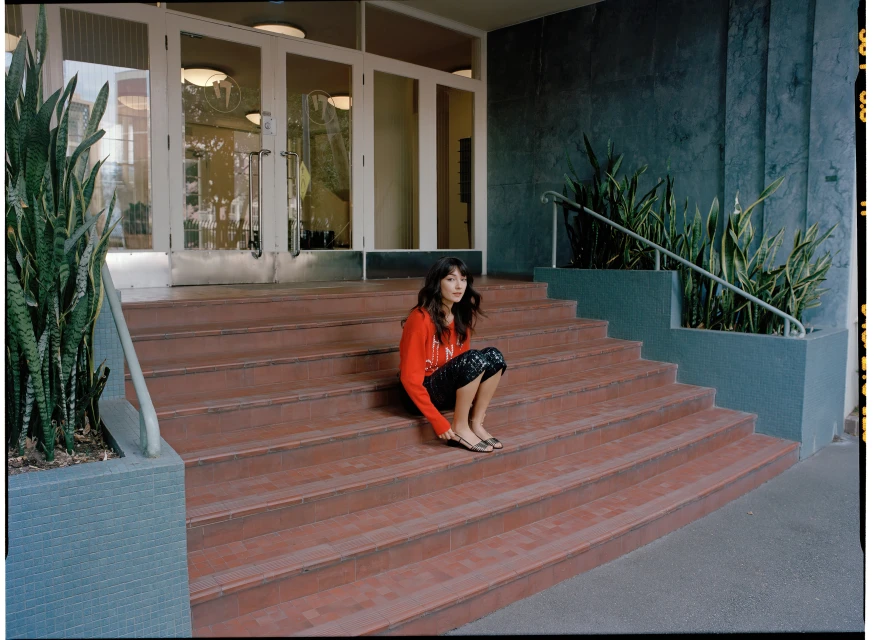 Adult woman sits on outdoor steps in a red sweater and patterned skirt. Lush plants and architectural doors in the background.