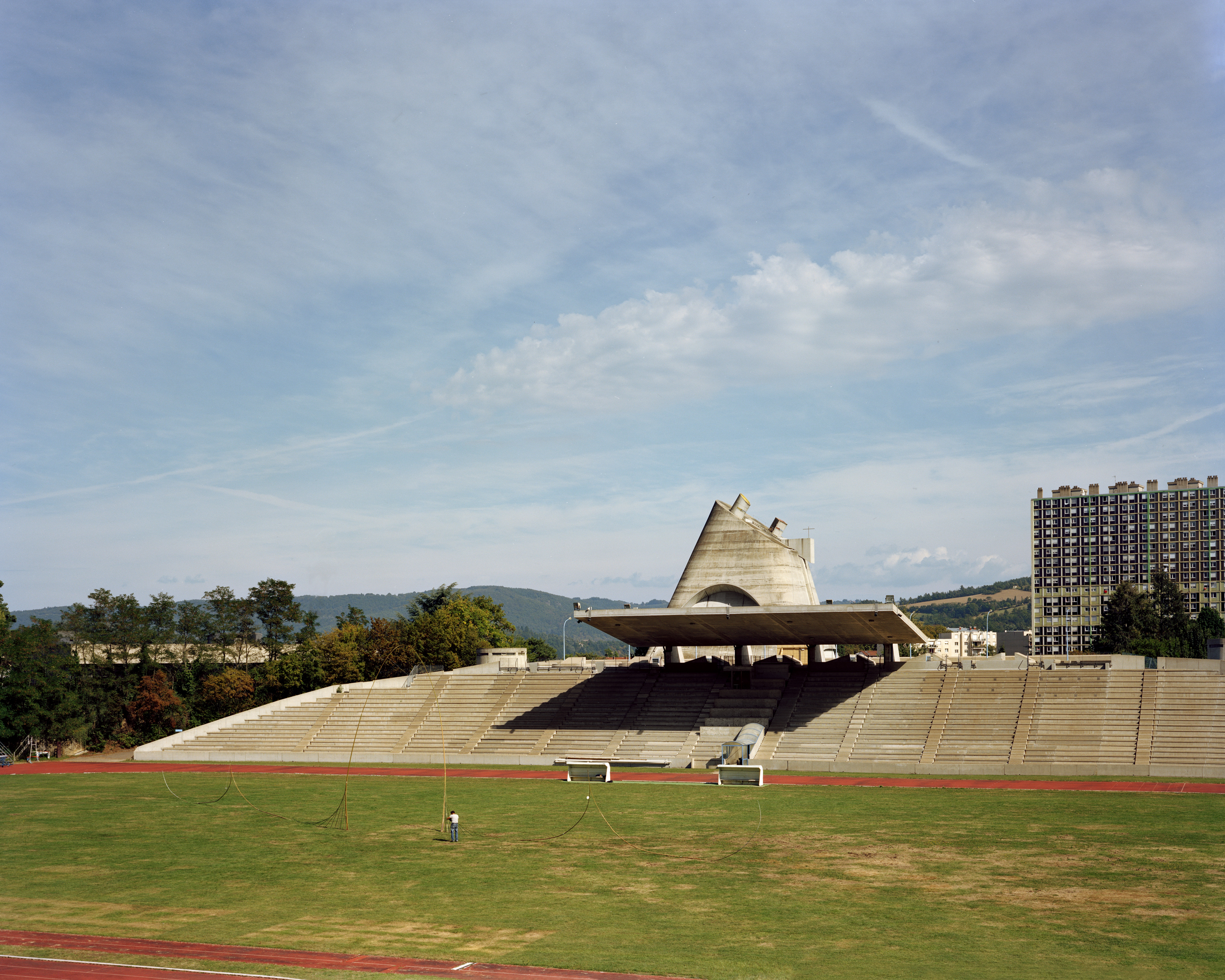 Wide shot of stadium seating under a unique concrete roof, with a running track, green field, and a distant apartment building.