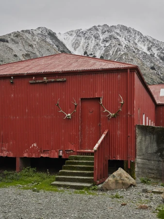 Bright red corrugated metal building with antlers mounted on the wall set against a snowy mountain background by Derek Henderson