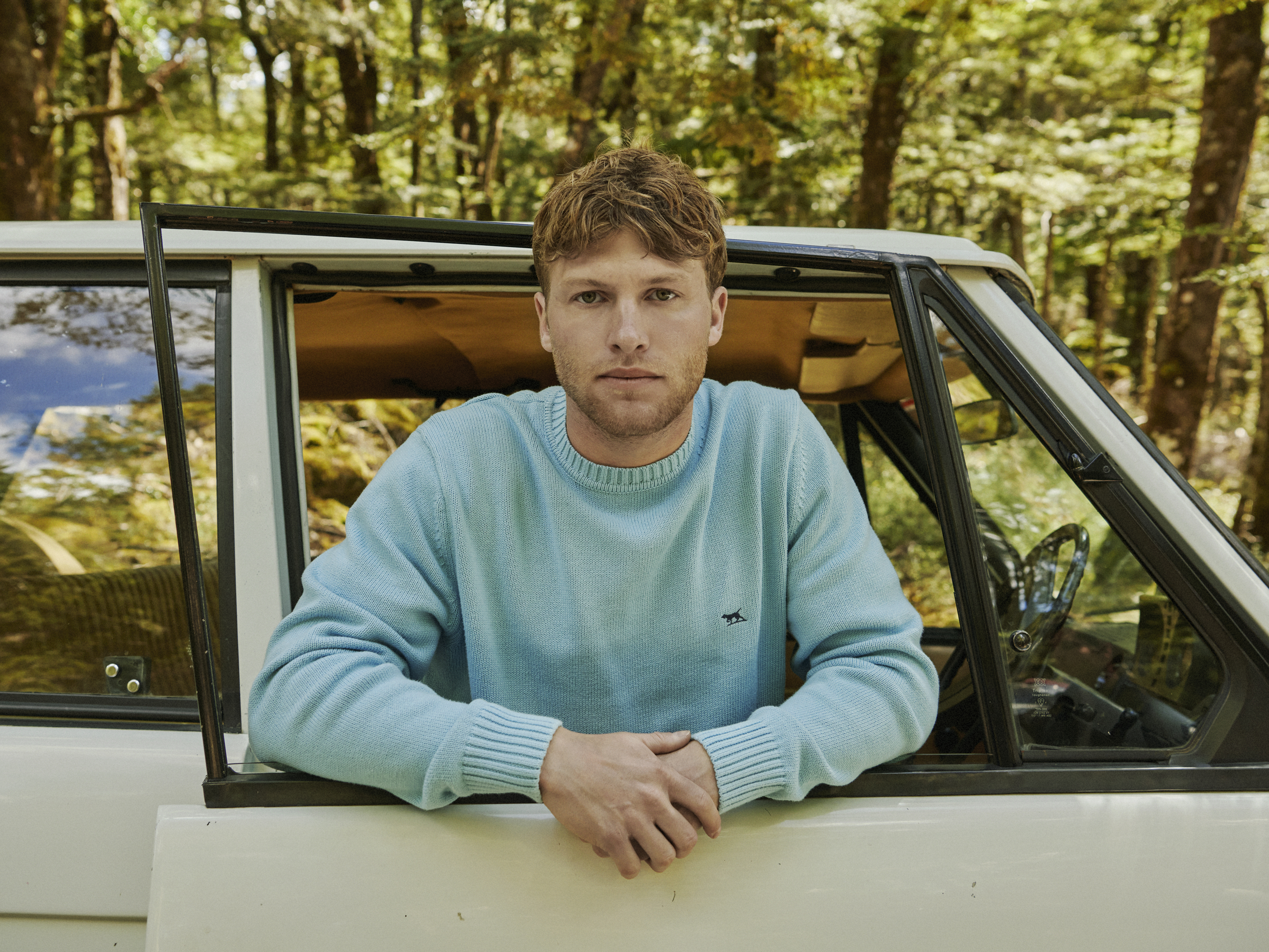A fair-skinned man with red hair wears a blue sweater and looks at the camera while leaning out the door of a vintage vehicle in a forest.