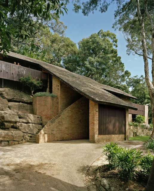 Exterior view of a brick and wood house nestled among green trees and large rock formations under a clear blue sky.