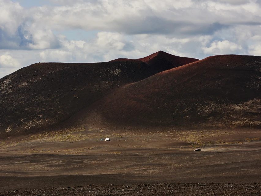 Volcanic landscape featuring a dark, red-toned mountain range with a cloudy sky. A distant car and tents dot the scene.