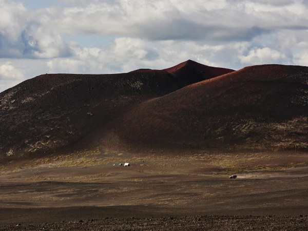 Volcanic landscape featuring a dark, red-toned mountain range with a cloudy sky. A distant car and tents dot the scene.