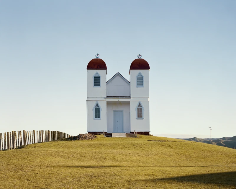 Architectural photograph of a small, white chapel with red domes on a grassy hill against a clear blue sky in a rural setting.