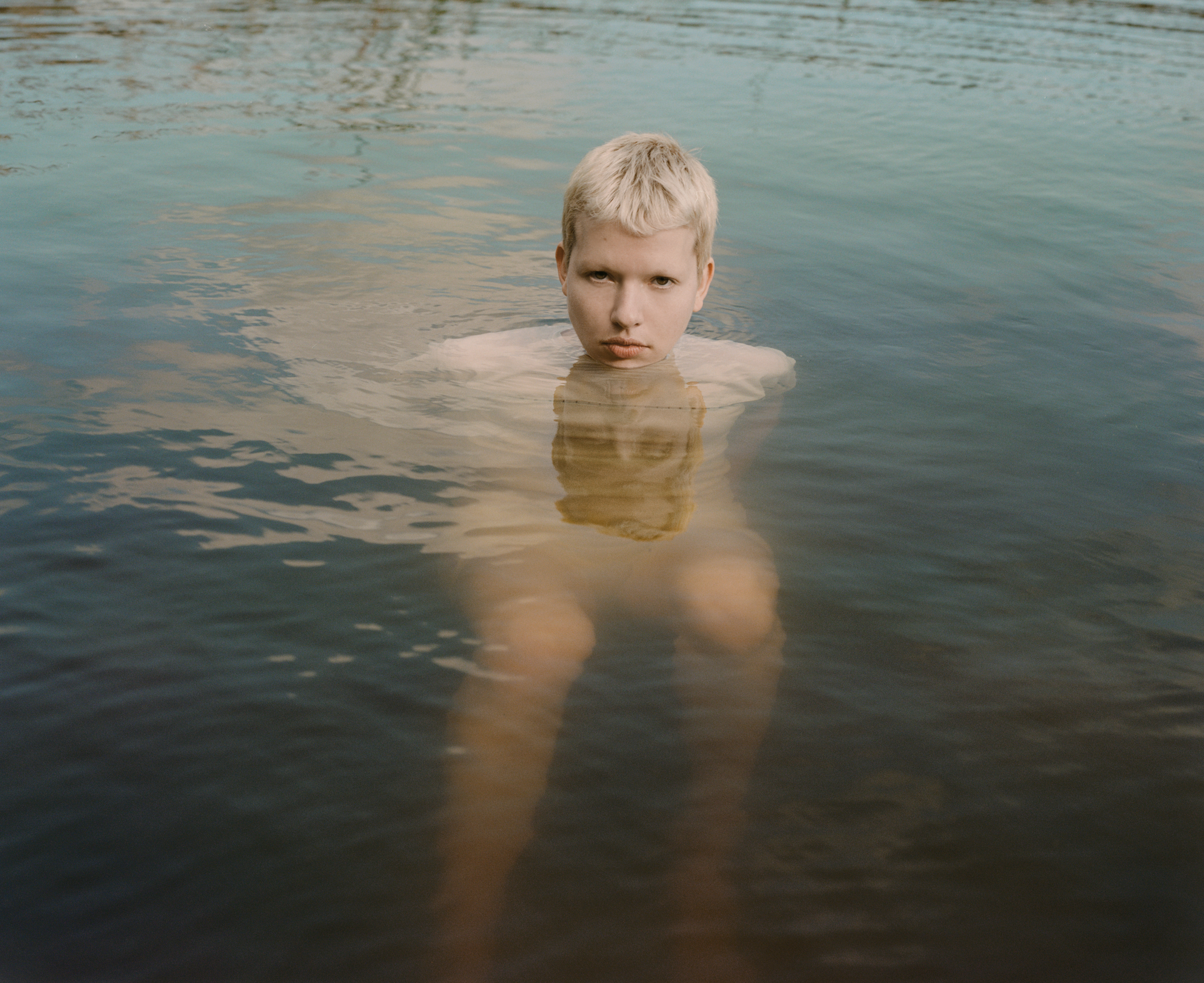 Fair-skinned person with short blonde hair stands waist-deep in water. The still water reflects the sky, creating a serene scene.