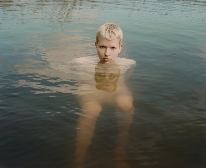 Fair-skinned person with short blonde hair stands waist-deep in water. The still water reflects the sky, creating a serene scene.