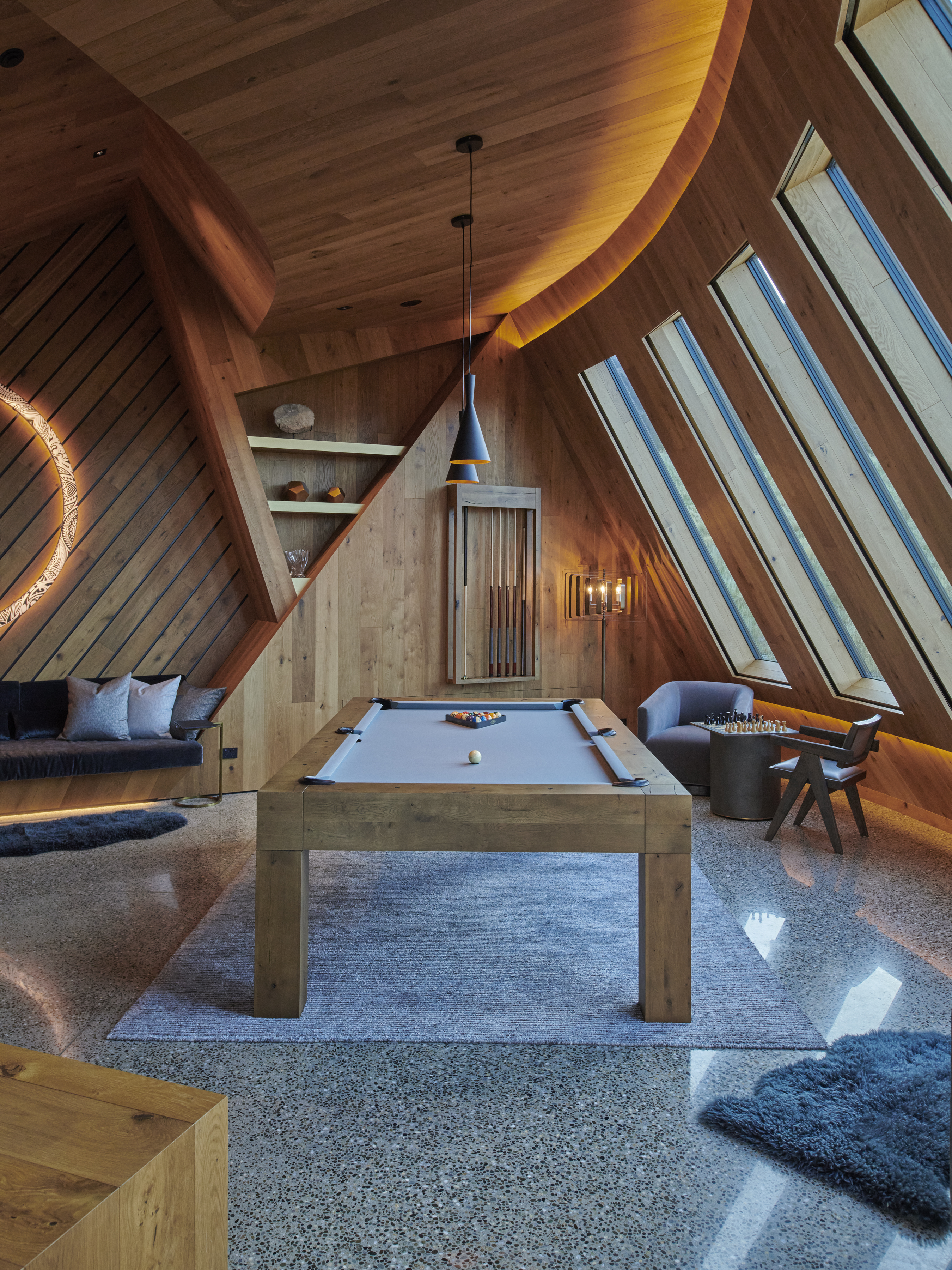 Indoor game room with pool table, wood paneled walls, angular architectural features, modern furniture and overhead lighting.