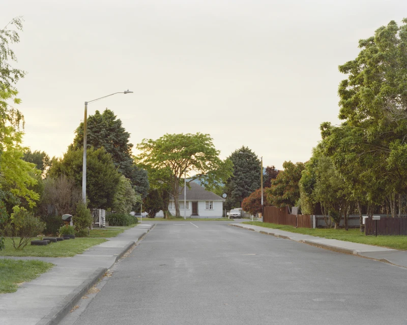 Quiet suburban street scene with a house, green trees, and a clear sky, capturing a sense of calm and neighborhood tranquility.