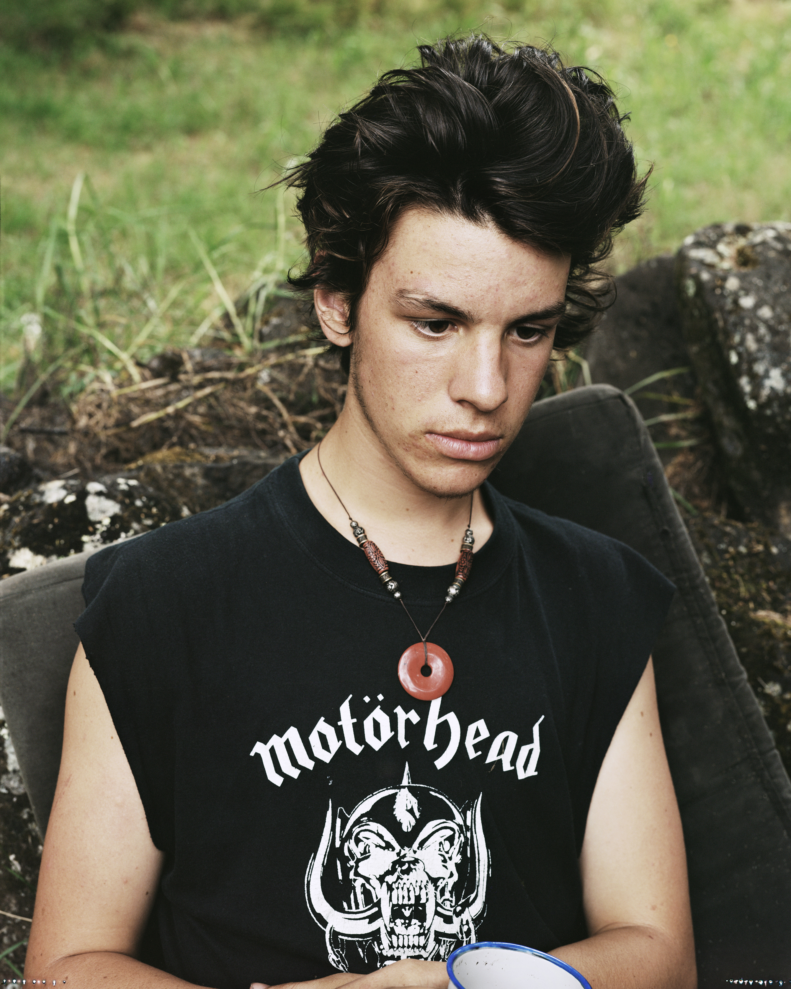 Portrait of a young adult male with dark hair wearing a black sleeveless shirt with the logo of Motorhead, sitting outdoors.