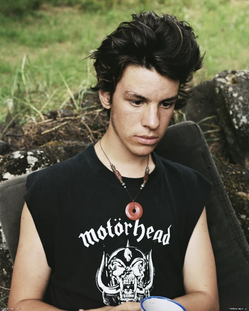 Portrait of a young adult male with dark hair wearing a black sleeveless shirt with the logo of Motorhead, sitting outdoors.