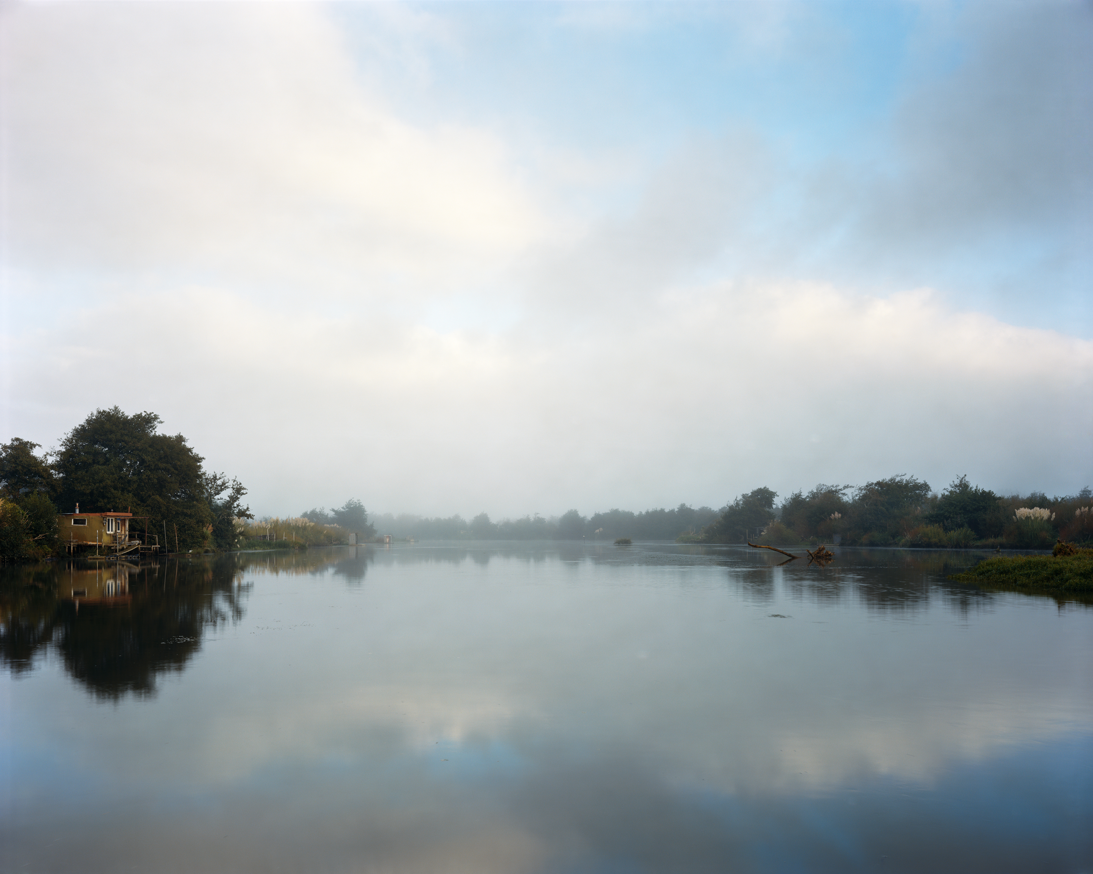 Foggy landscape photo features a serene lake reflecting a small structure surrounded by trees, beneath an overcast sky with soft light.