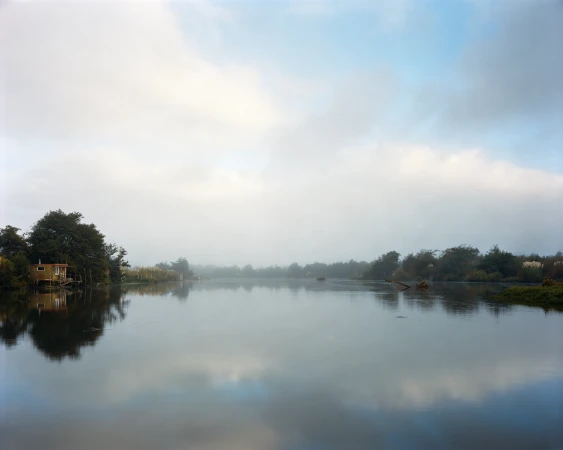 Foggy landscape photo features a serene lake reflecting a small structure surrounded by trees, beneath an overcast sky with soft light.