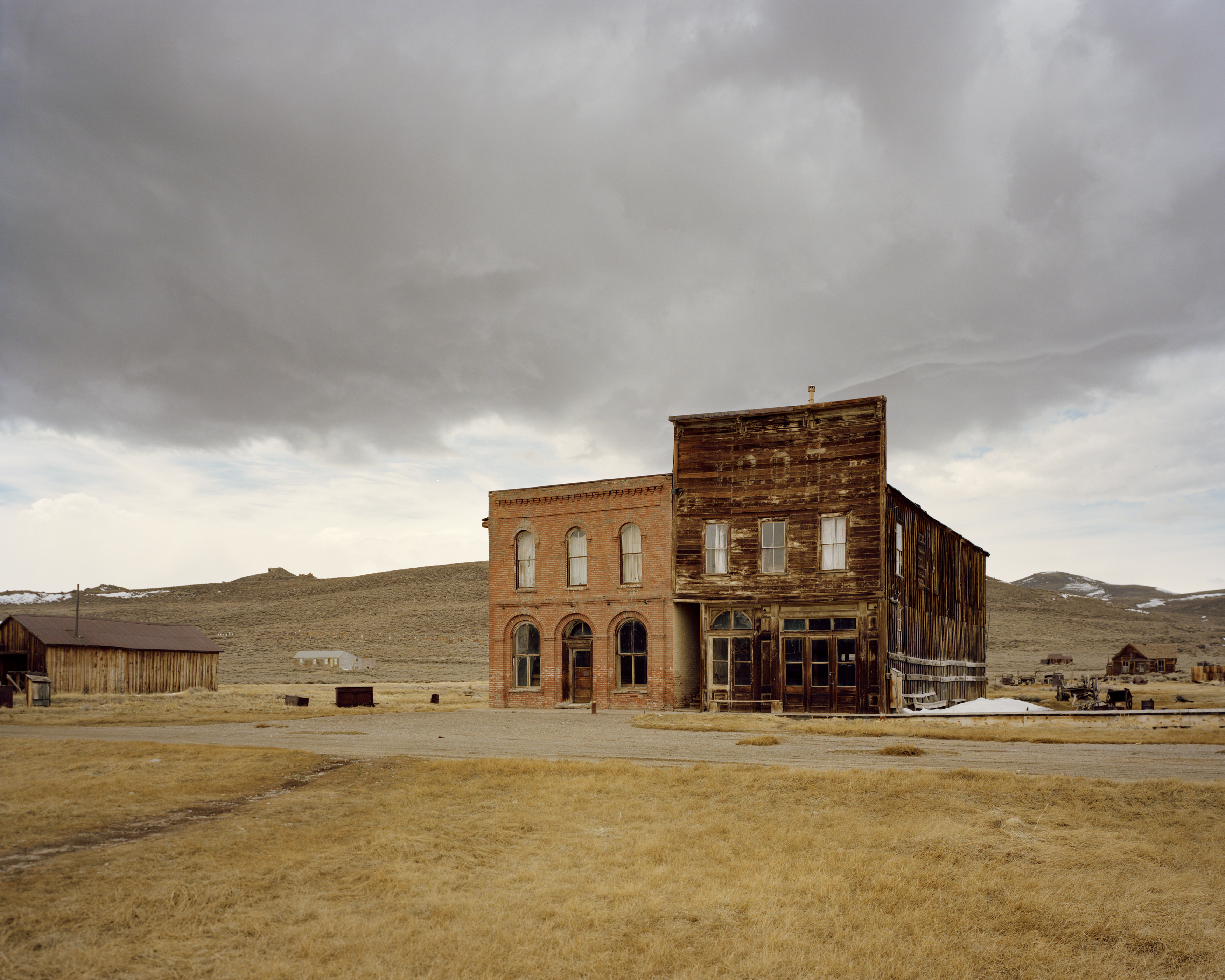 Old brick and wood buildings stand in a deserted town under a cloudy sky in an open field with hills in the background.
