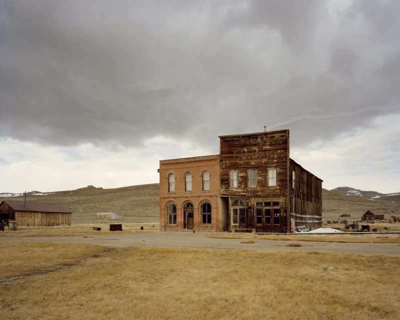Old brick and wood buildings stand in a deserted town under a cloudy sky in an open field with hills in the background.