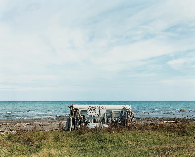 Dilapidated trailer on a beach with ocean and sky background, covered in branches and foliage, creating an unusual and surreal scene.