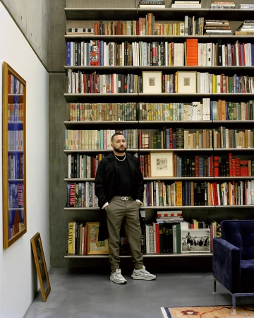 Man standing in front of a tall, fully stocked floor-to-ceiling bookshelf in a modern room, photographed by Derek Henderson