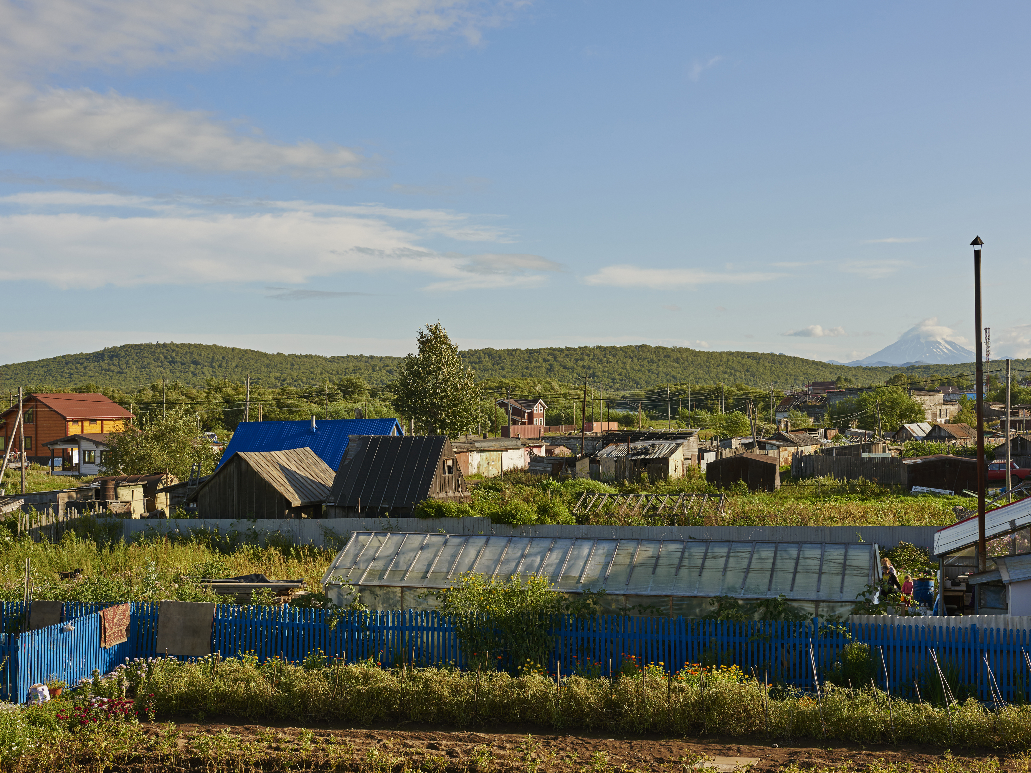 A rural village scene with houses, greenhouses, and a blue fence in a grassy field under a blue sky with distant mountain views.