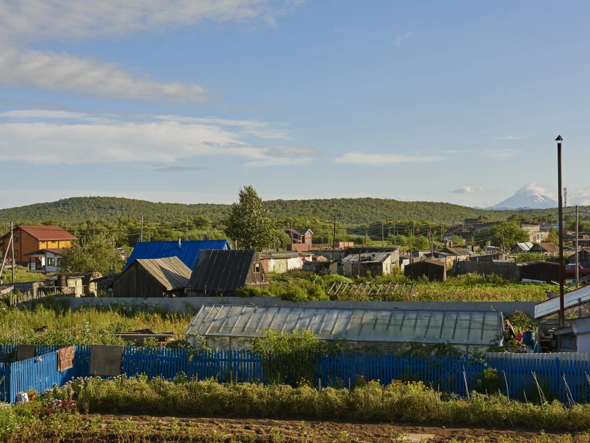 A rural village scene with houses, greenhouses, and a blue fence in a grassy field under a blue sky with distant mountain views.