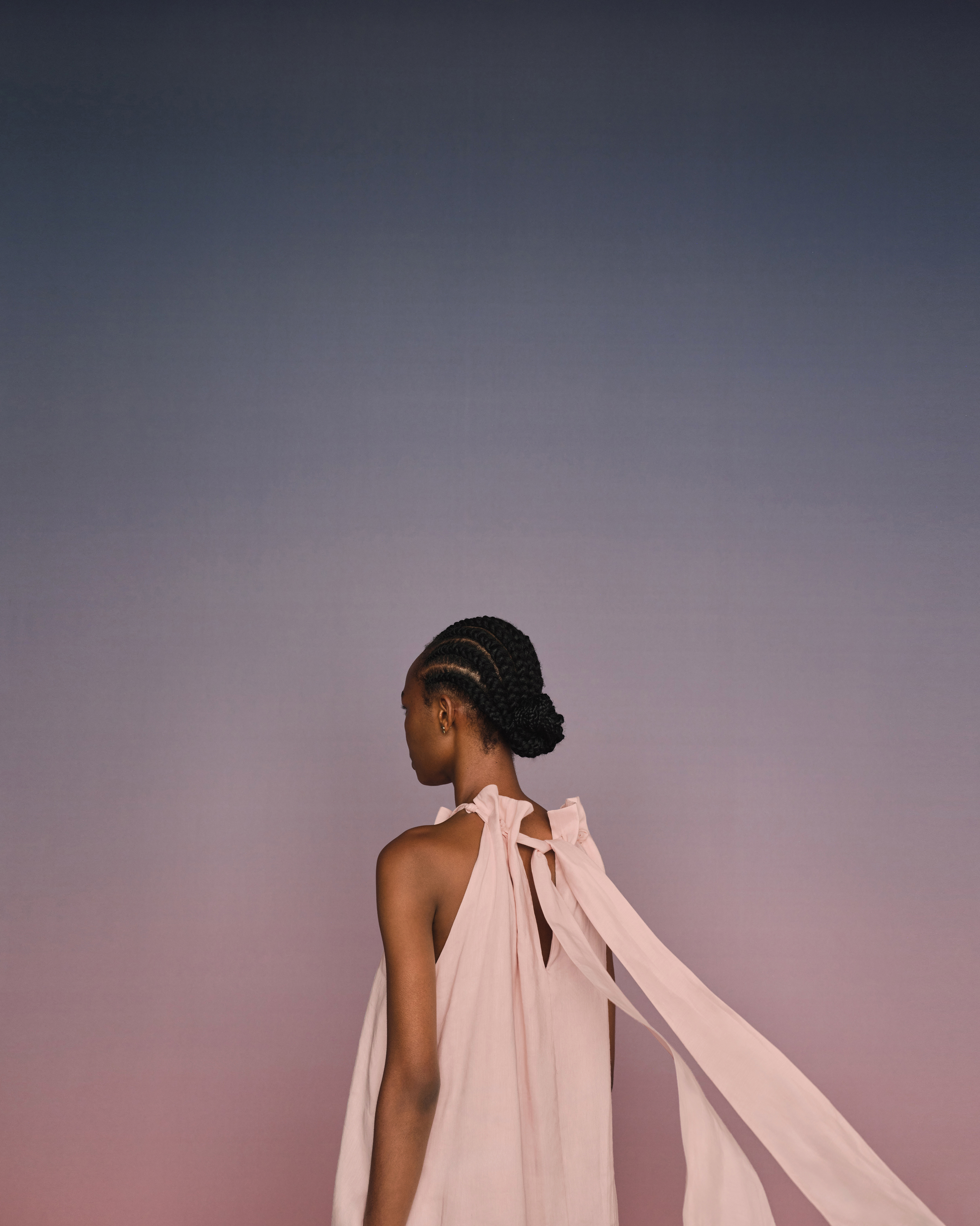 Model seen from behind wearing a pink sleeveless dress with flowing ribbons against a gradient background