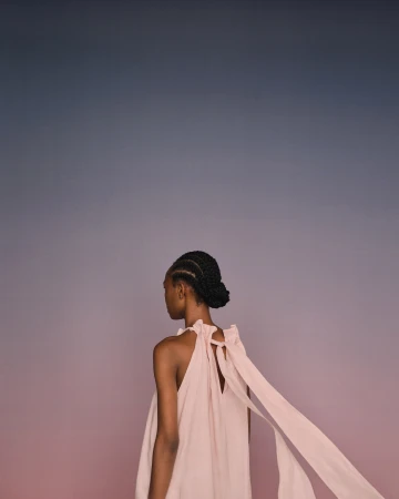 Model seen from behind wearing a pink sleeveless dress with flowing ribbons against a gradient background