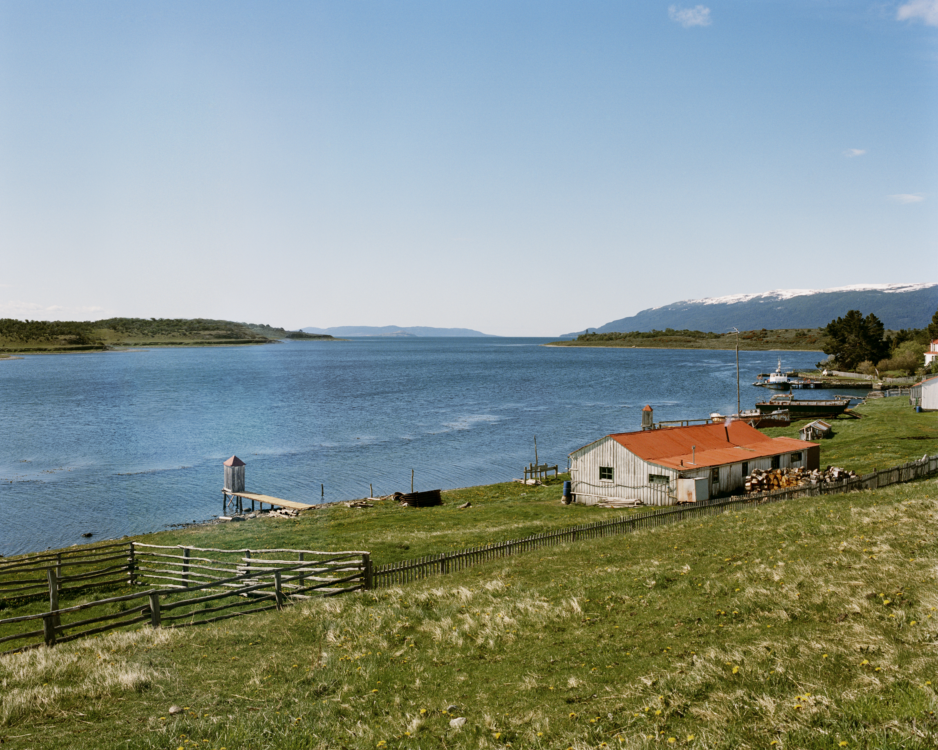 Scenic daytime view of a lake, a wooden house with a red roof, green meadow with a fence, and distant mountains with snow-capped peaks.