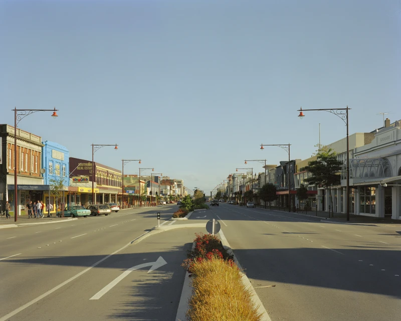 Urban thoroughfare with multiple cars and pedestrians on the sidewalks, buildings on either side, beneath a clear blue sky.