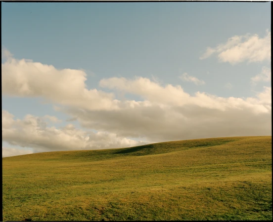 Rolling grassy green meadow beneath a vast pale blue sky filled with soft white clouds photographed by Derek Henderson