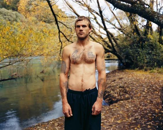 Shirtless tattooed man standing by a riverbank in a wooded area with fallen autumn leaves